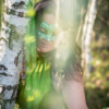 Iris lächelt mit grüner Glitzermaske hinter Birkenstamm hervor