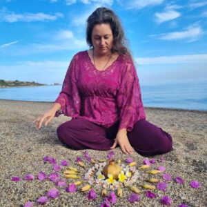 Iris sitzt vor einem Mandala aus Steinen und Blüten am Strand, im Hintergrund das Meer