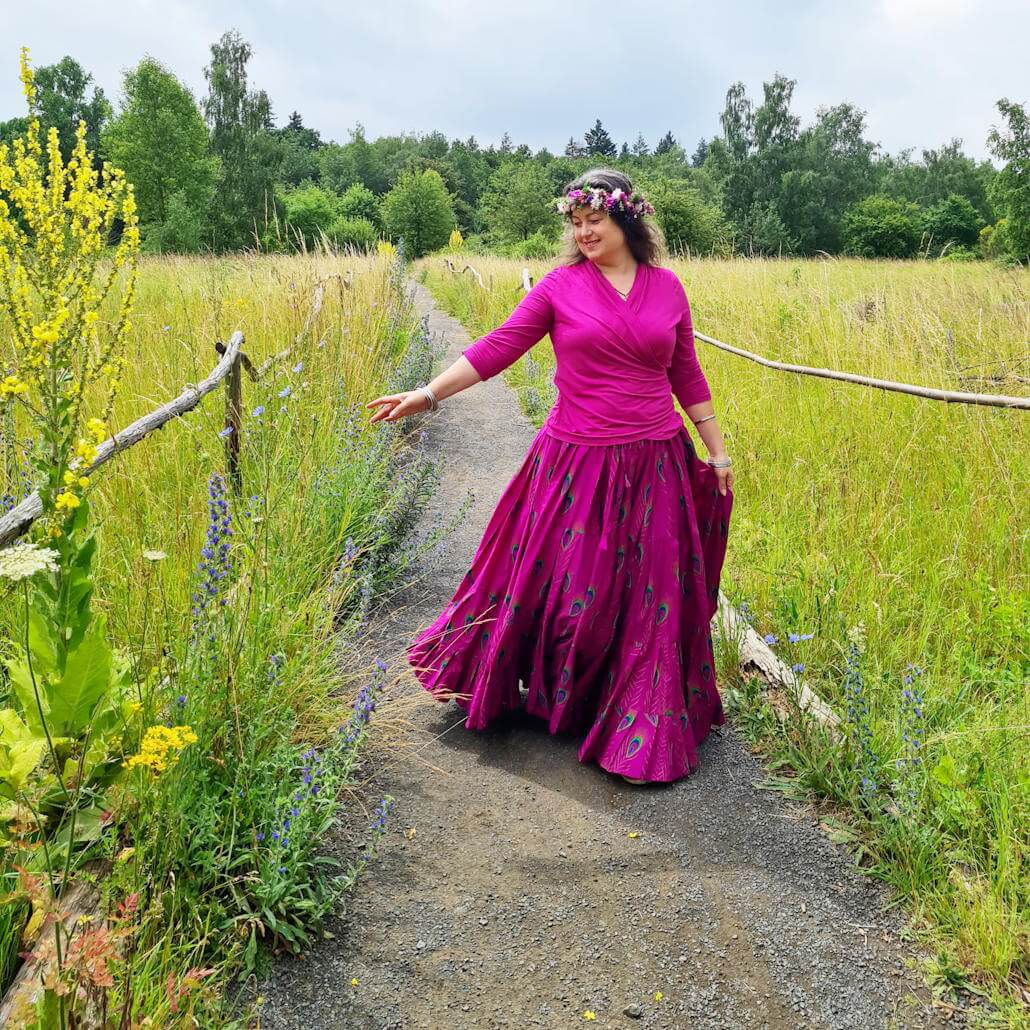 Iris läuft mit pink-farbenem Kleid und Blütenkranz auf dem Kopf auf einem Feldweg in einer Wiese